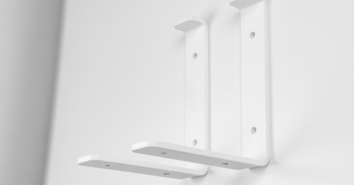 Two plain white shelf brackets sit on a plain white surface. The brackets have an L-shape with four screw holes each.
