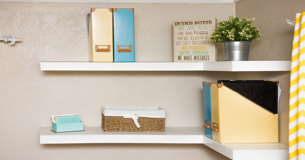 Two white floating corner shelves on a beige wall. The shelves contain home decor items, books, and a plant.