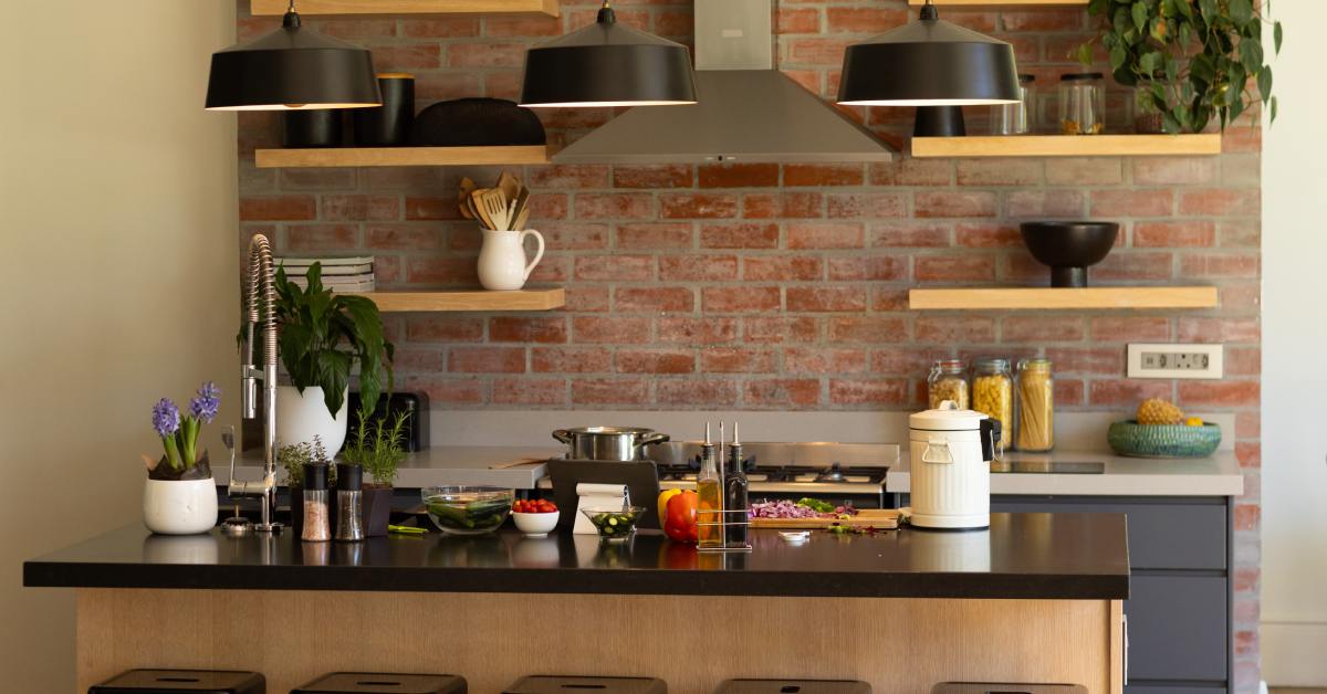 A kitchen with an exposed brick wall behind the stove and primary work surface. There are wooden floating shelves.