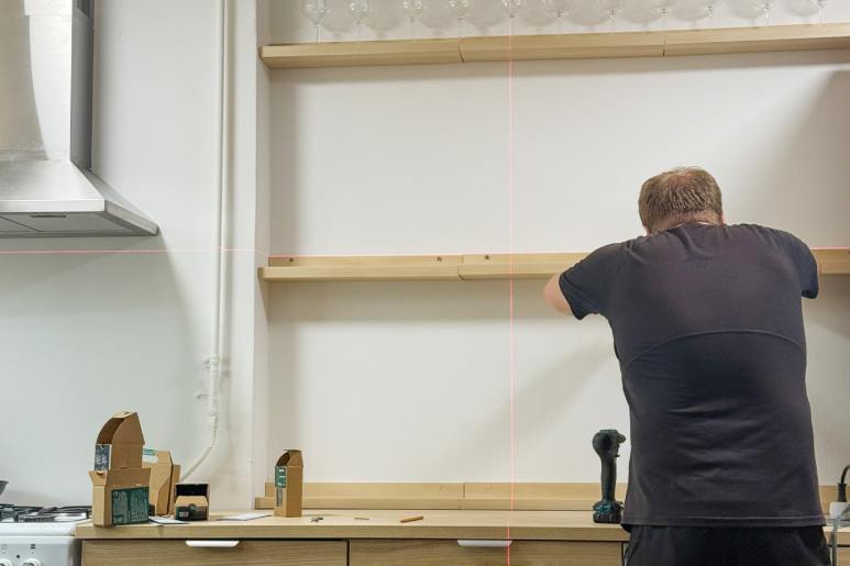A person stands at the kitchen counter as they lean over to install floating shelves. They hold up the bottom shelf.