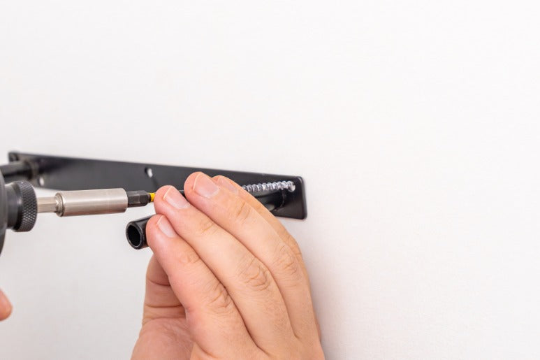 A close-up of a person installing a black shelf bracket on a white wall. They use a screw gun to mount the nail.