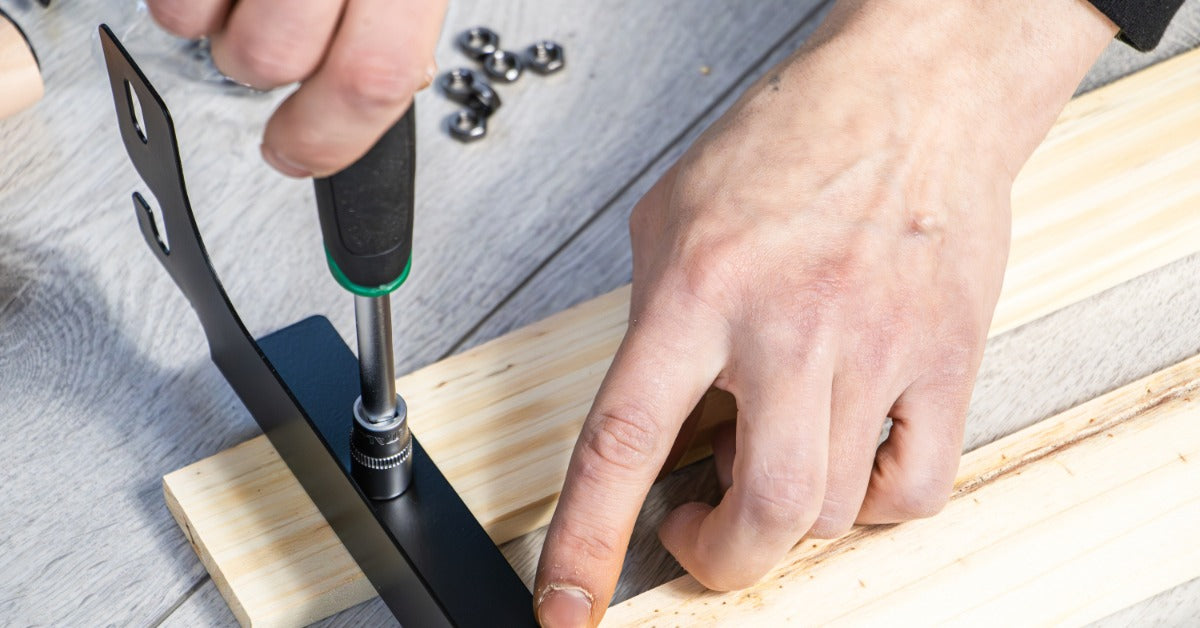 A close-up of someone's hands installing a black bracket to a piece of wood. They're using a screwdriver.