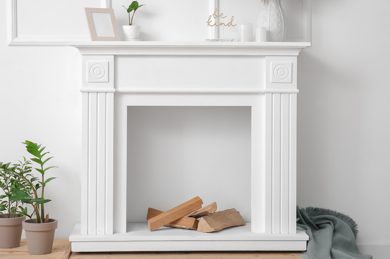 A white faux fireplace mantel on a white wall holds a picture frame, small plant, candles, and a large vase with branches.