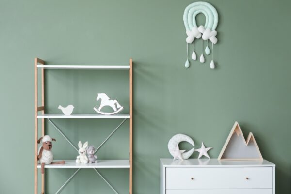 A baby nursery with a green wall, a bookshelf with toys, and a white chest of drawers with neutral-colored decorations.