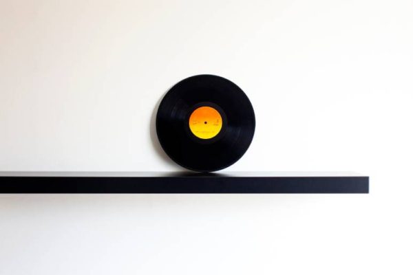 One black floating shelf mounted against a white wall holds one single black and yellow vinyl phonograph record.