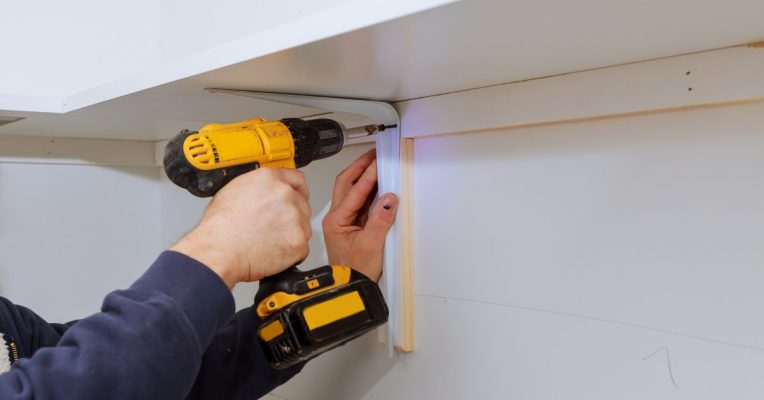 A person holding a cordless power drill. They're installing a screw onto a bracket under a white corner shelf.