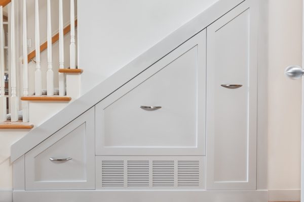 A view of a white staircase with wooden accents and the wall and space underneath it which includes pullout cabinets.