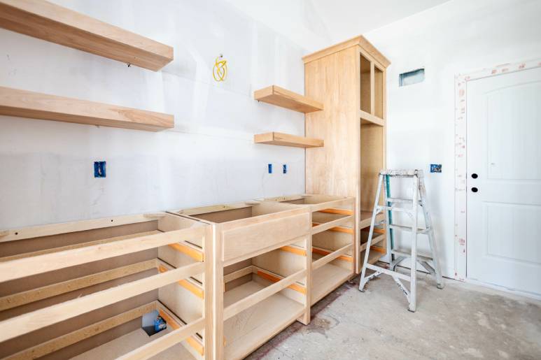 A room under construction featuring floating wooden shelves and cabinets. A step ladder is propped by the door.