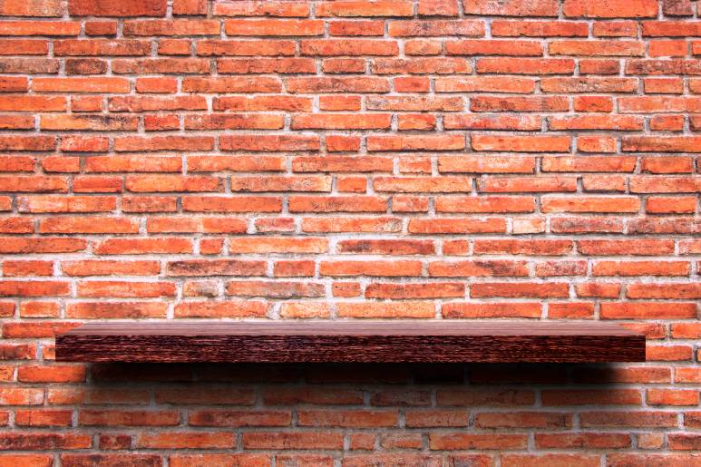 A single floating shelf made of darkly colored wood is installed onto a red brick wall. The shelf is empty.