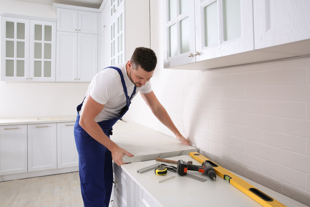 An individual wearing blue work overalls holds the surface of a countertop as he installs it in position.