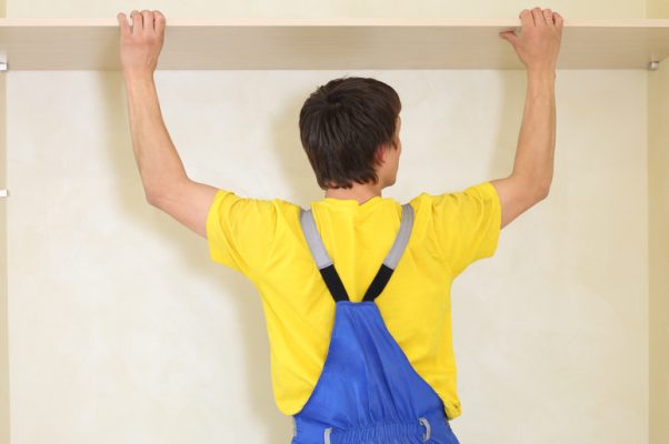 An individual in a yellow shirt and blue overalls raises their arms as they try to fit a shelf into a wall.