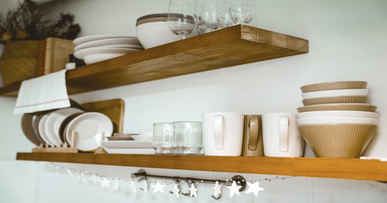 Two floating shelves mounted to the wall of a kitchen. The shelves hold glasses and brown and white bowls, plates, and cups.